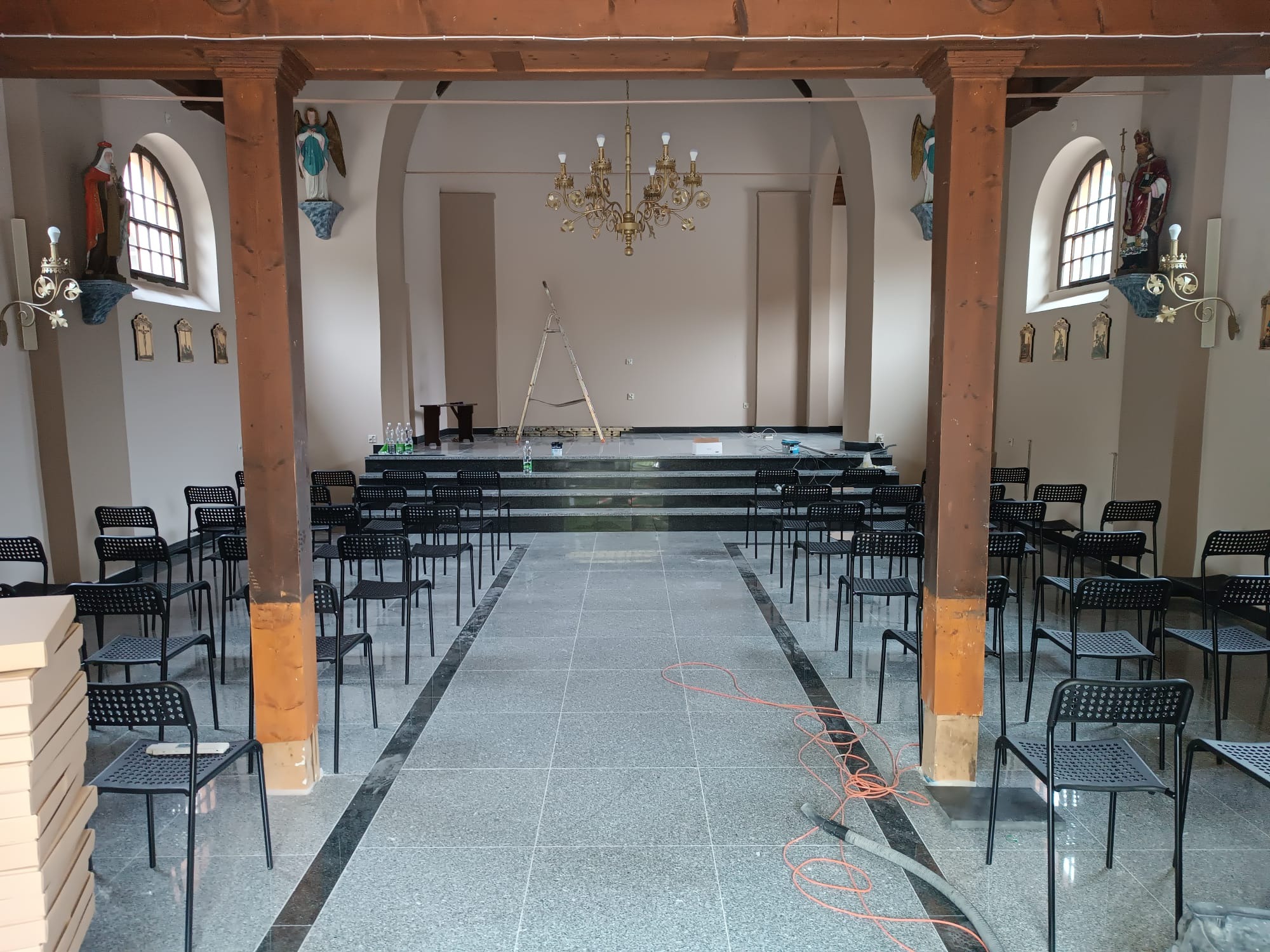 Wnętrze kościoła z nową posadzką z szarego kamienia, rzędy czarnych krzeseł, drewniane kolumny i złoty żyrandol, drabina oparta o podwyższenie o kamiennych schodach, widoczne ślady prac...