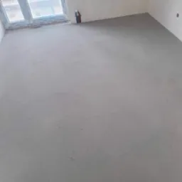 Świeżo wylana, szara posadzka w pustym pomieszczeniu z oknem, widoczne wystające rurki instalacji.