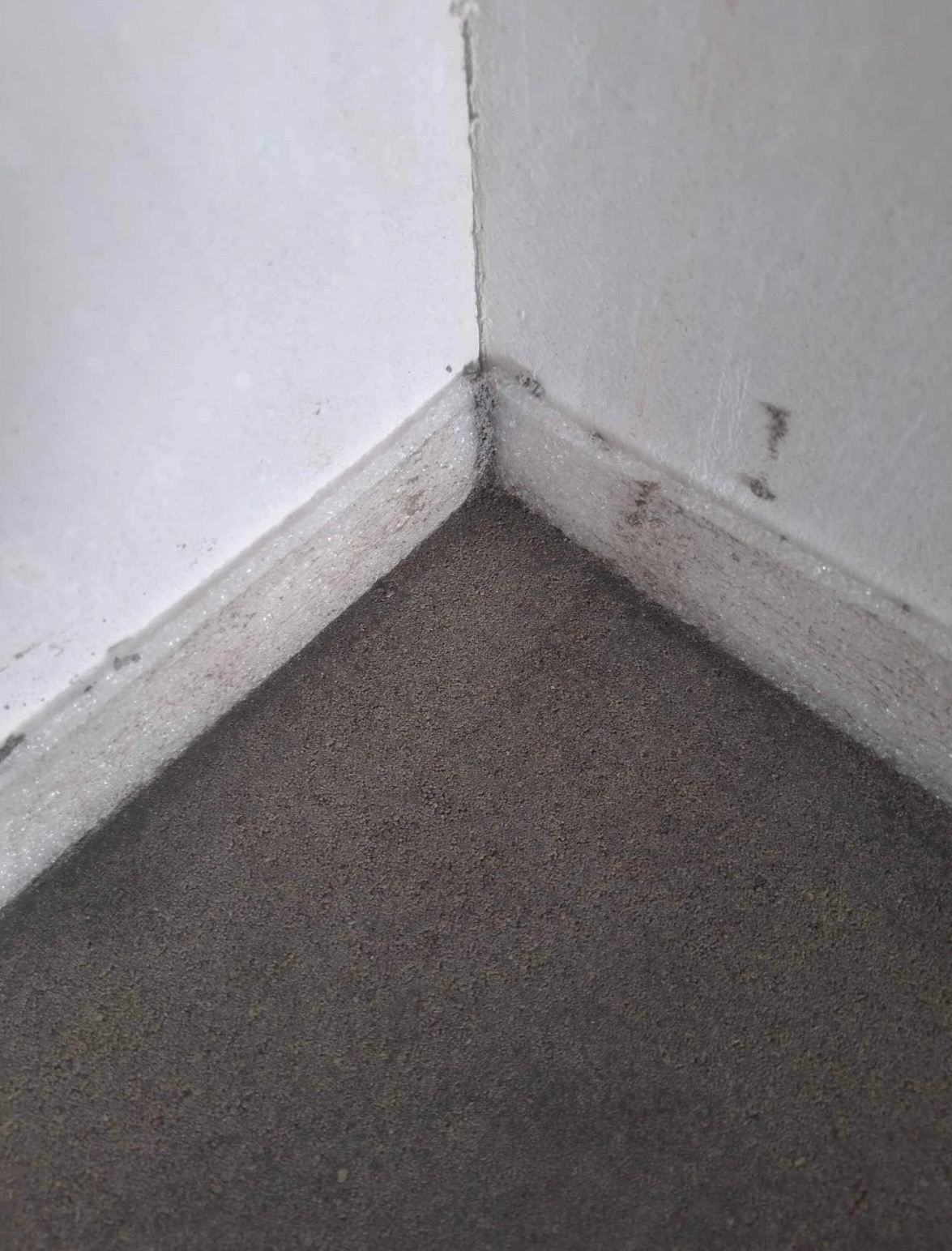 Świeżo wylany, ciemnoszary jastrych przy ścianie, zabezpieczony folią przy krawędziach, widok z góry w rogu pomieszczenia.