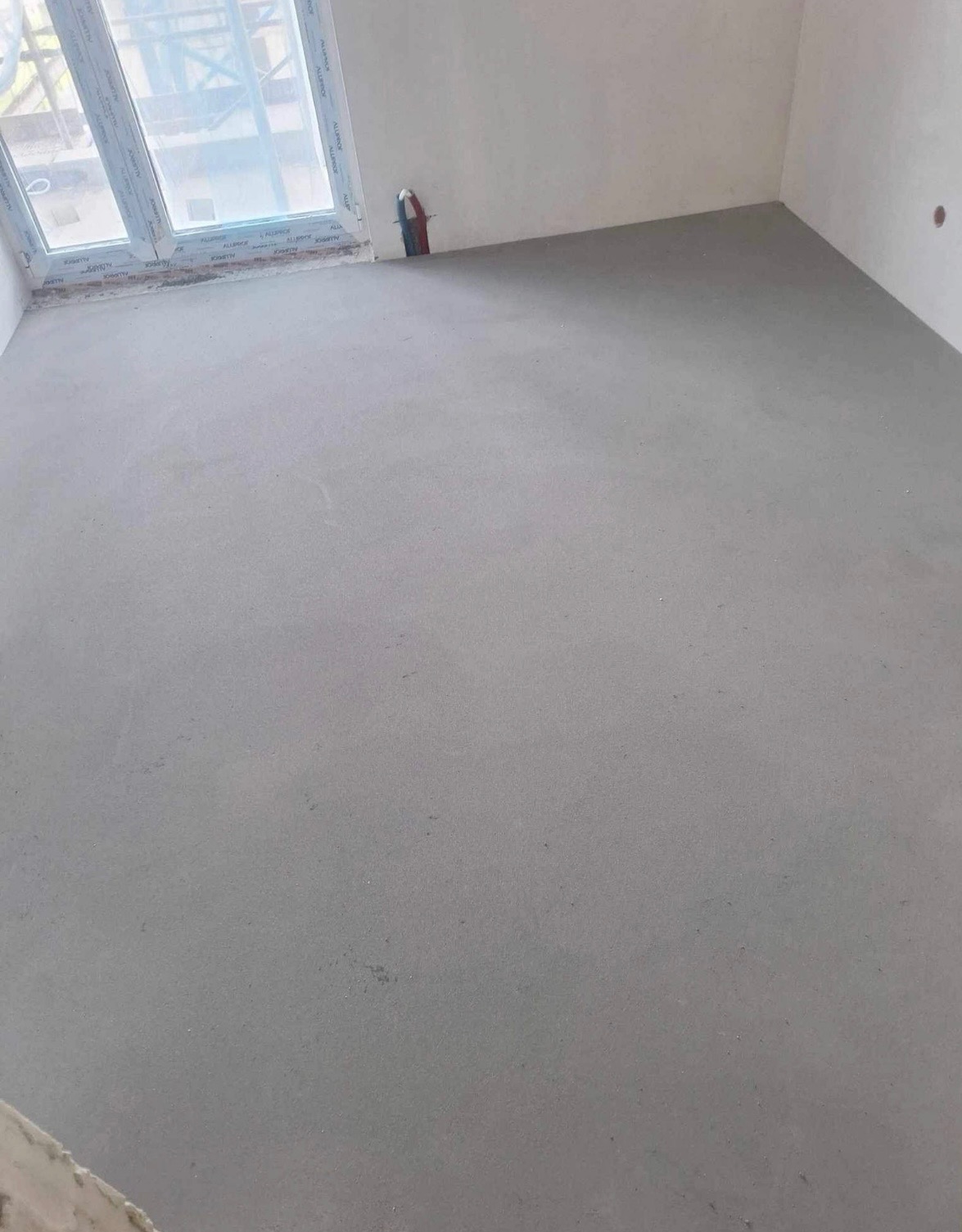 Świeżo wylana, szara posadzka w pustym pomieszczeniu z oknem, widoczne wystające rurki instalacji.