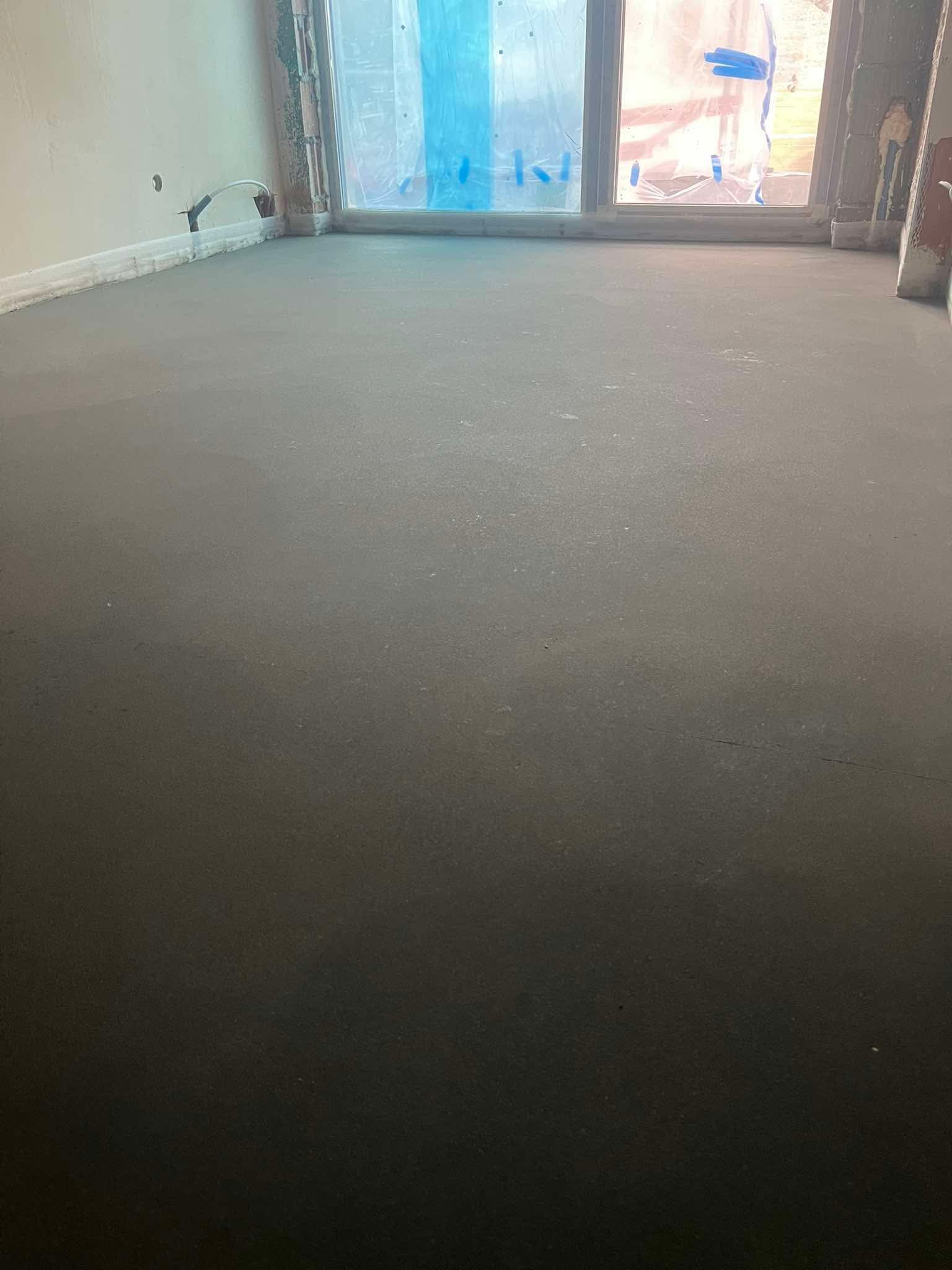 Świeżo wylana, gładka posadzka w jasnym pomieszczeniu z oknem zabezpieczonym folią. Widoczne listwy przypodłogowe i fragment ściany w surowym stanie.