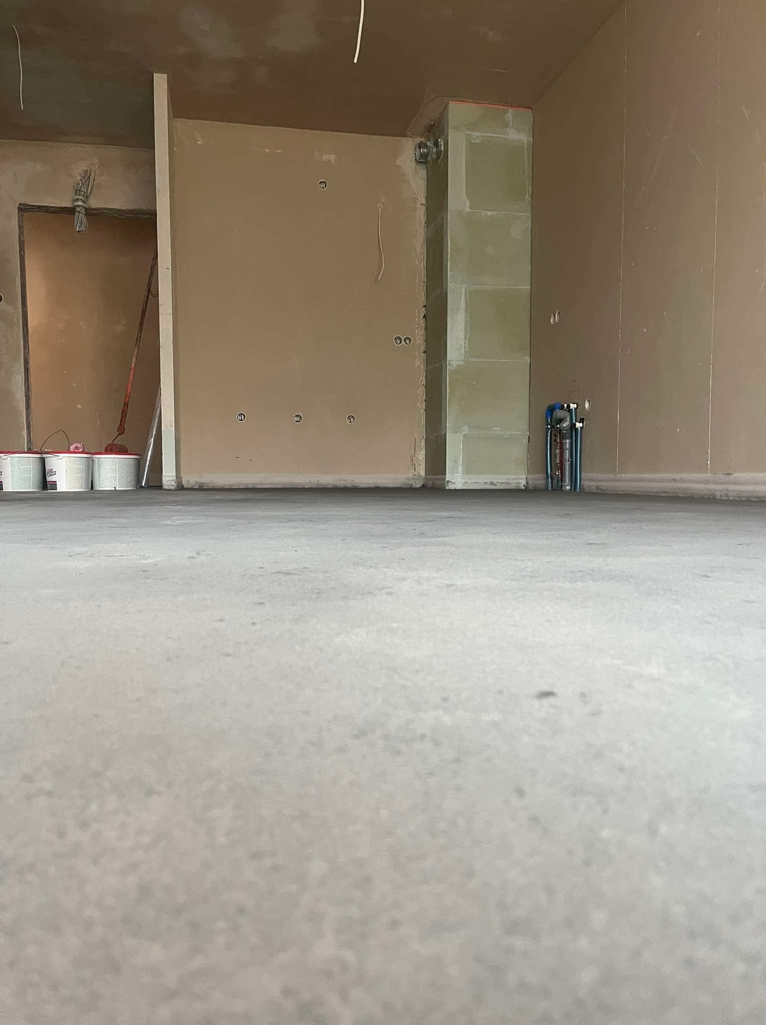 Wnętrze w trakcie prac wykończeniowych: gładka, jasna posadzka jastrychowa, ściany przygotowane do malowania, widoczne instalacje i materiały budowlane.