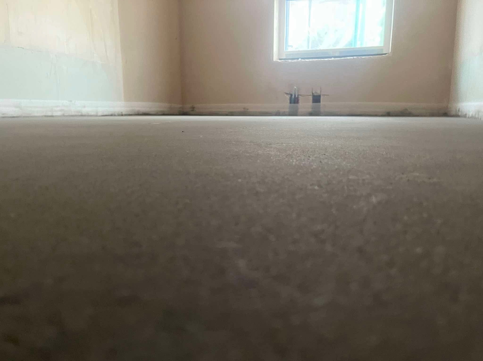 Wylana, szara posadzka jastrychowa w pokoju z oknem. Widoczne wystające rurki instalacji. Ściany w jasnym odcieniu beżu.