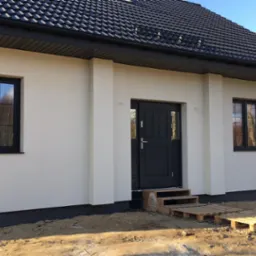 Nowoczesny dom jednorodzinny z kremową elewacją, czarną stolarką okienną i drzwiami, oraz ciemną dachówką. Schody wejściowe tymczasowo wykonane z palet. Widoczne przygotowane podłoże wokół budynku.