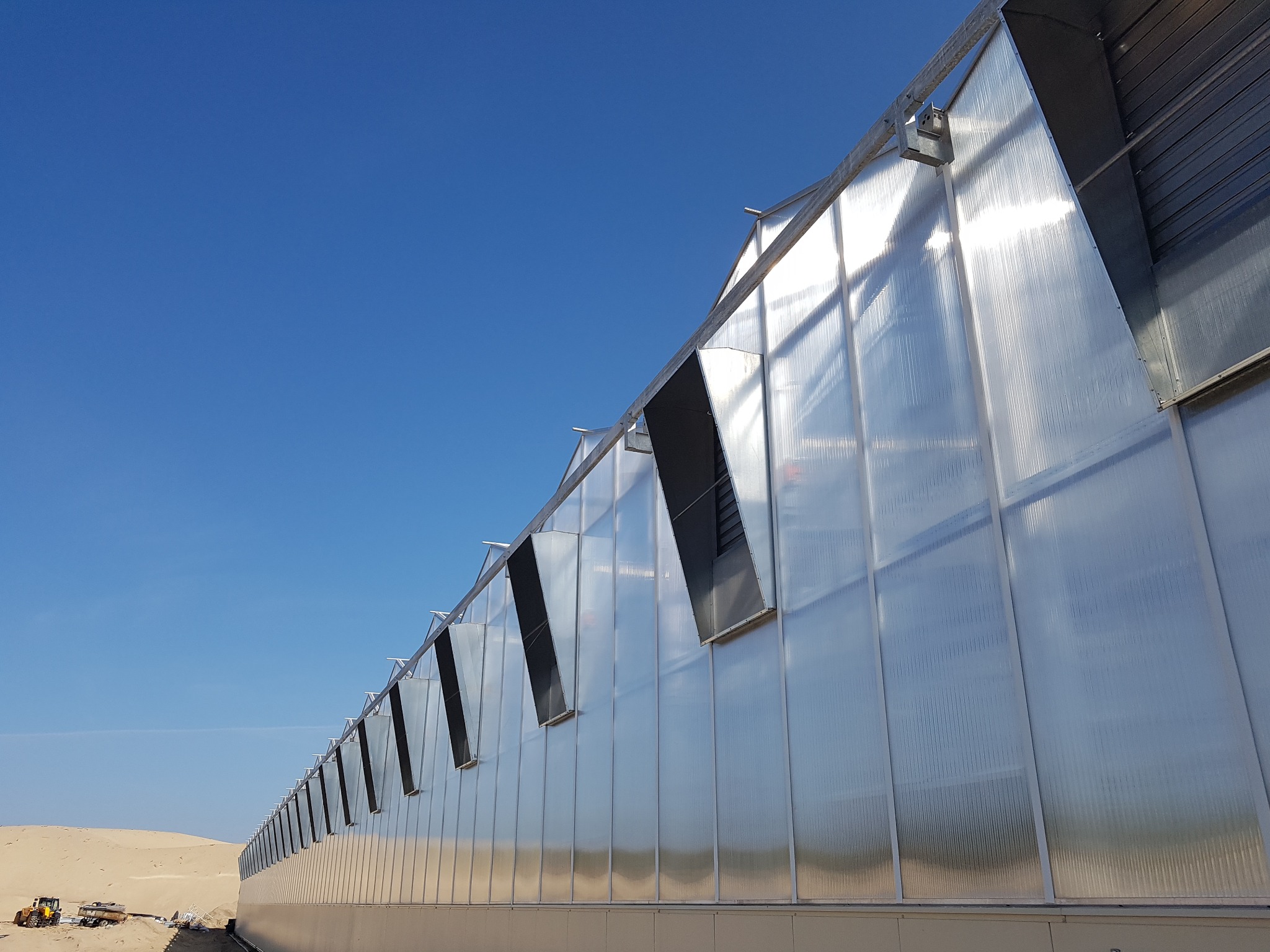 Zewnętrzny widok szklarni z systemem wentylacji w postaci otwieranych paneli, na tle piaszczystego terenu i błękitnego nieba.