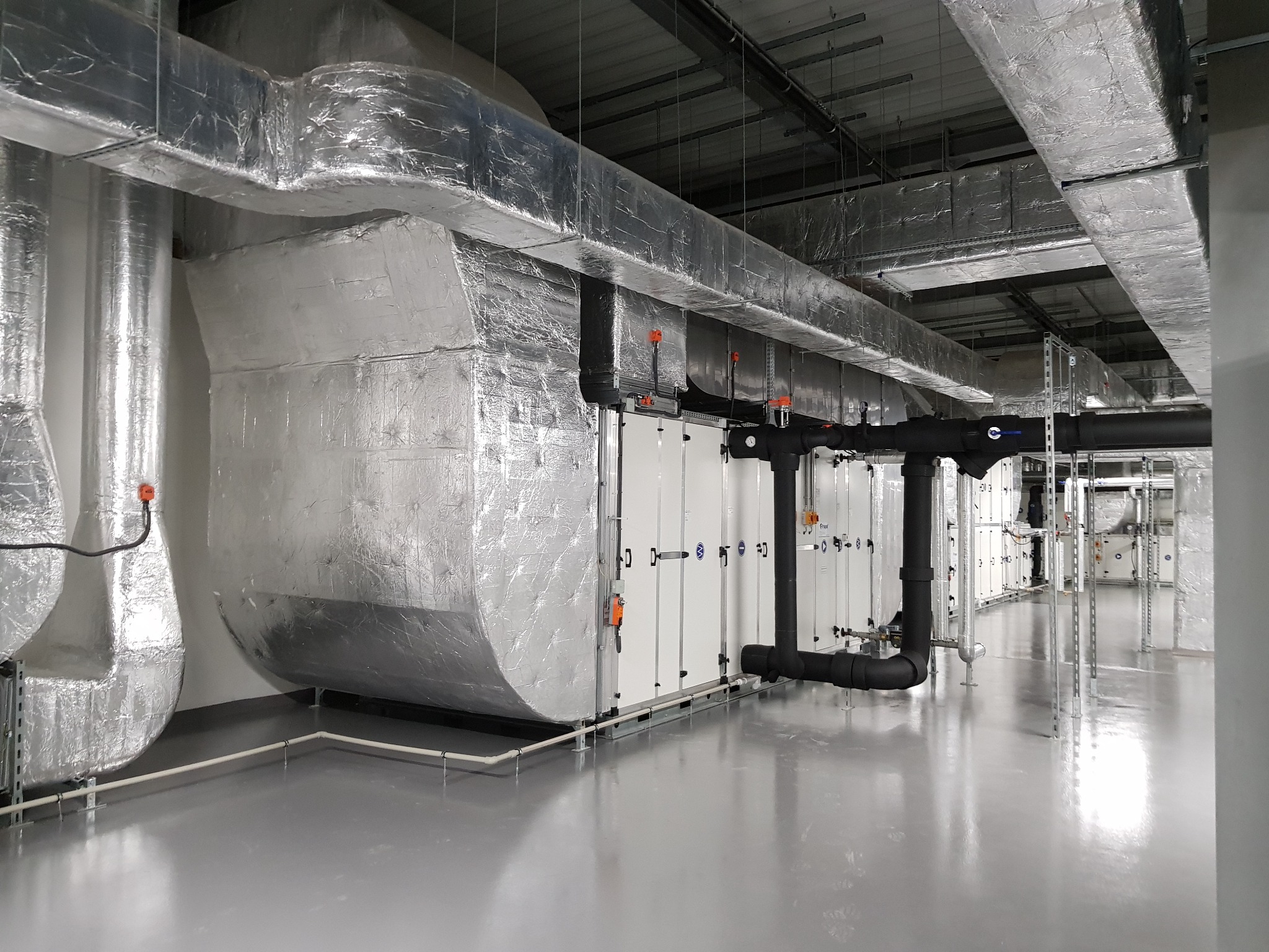 Widok instalacji wentylacyjnej z izolowanymi kanałami i centralami wentylacyjnymi w przemysłowym budynku, z czarnymi rurami i szarą podłogą. Widoczne elementy mocujące i przewody.