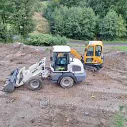 Dwa urządzenia budowlane na placu budowy: biała ładowarka kołowa KMM i żółta minikoparka Robex R16-9AK, widoczne ślady opon na ziemi, w tle drzewa i pochmurne niebo.