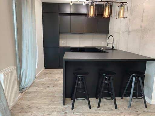 Nowoczesna, czarna kuchnia z wyspą i trzema hokerami, oświetlona wiszącymi lampami z metalowymi kloszami, z widocznymi elementami grzejnika i zasłony.