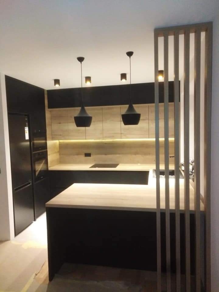 Nowoczesna kuchnia z czarnymi frontami i drewnianymi blatami, oświetlenie punktowe i wiszące lampy, oddzielona ażurową ścianką z drewnianych listew.