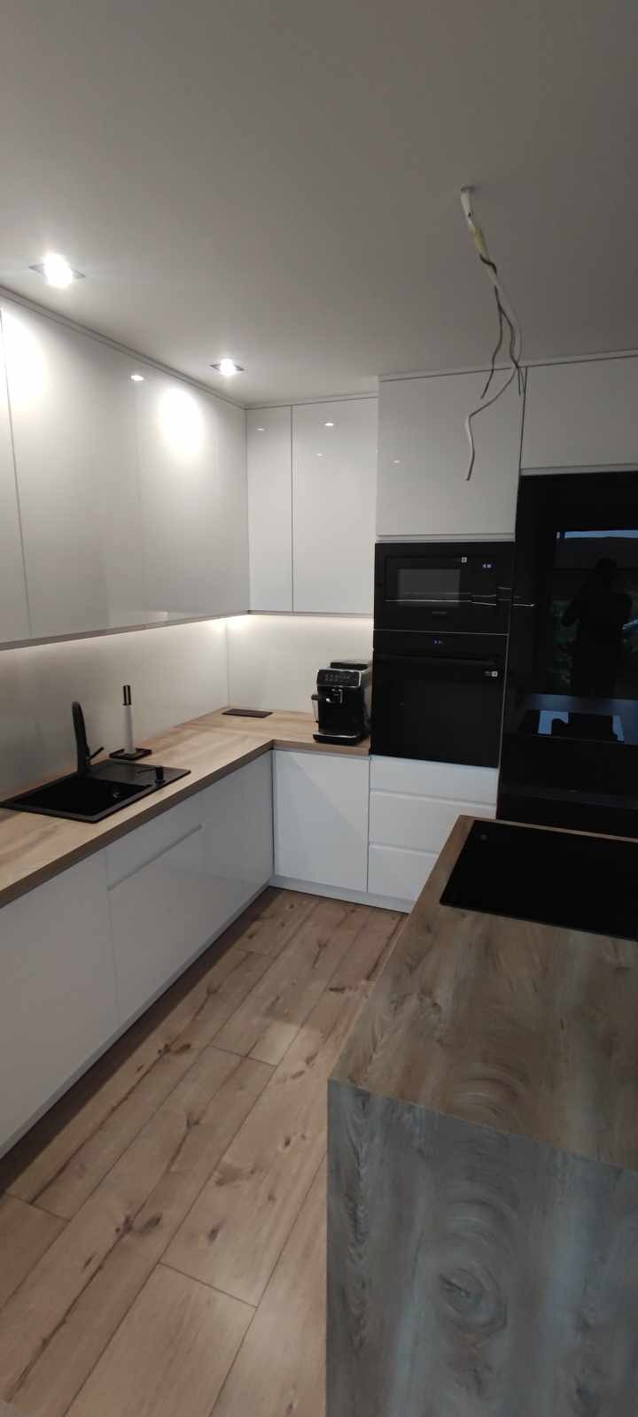 Nowoczesna kuchnia z białymi, lakierowanymi frontami, drewnianym blatem i czarnym sprzętem AGD, widoczne niedokończone podłączenie elektryczne.