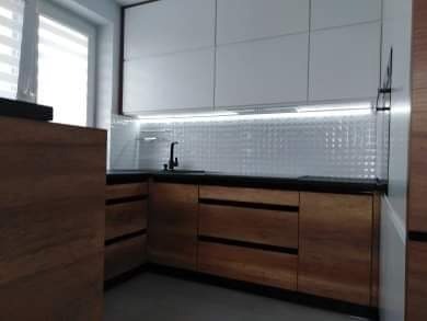 Nowoczesna kuchnia: białe górne szafki, drewniane dolne z poziomymi uchwytami, czarny blat i biała mozaika na ścianie, oświetlenie LED pod szafkami.