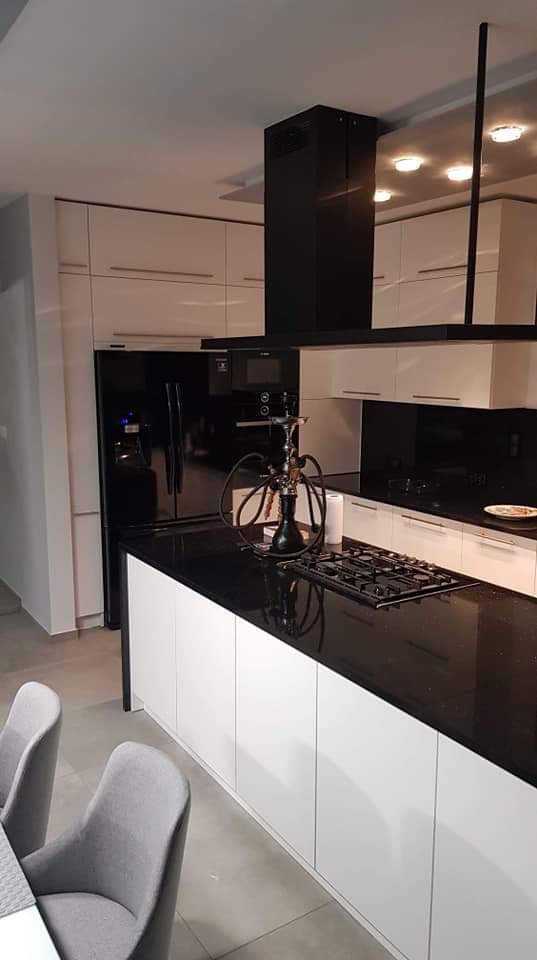 Nowoczesna kuchnia z białymi szafkami, czarnym blatem i okapem. Na blacie stoi szisza obok płyty grzewczej. Widoczne szare krzesła.