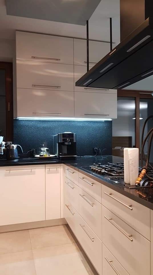 Nowoczesna kuchnia z białymi szafkami, czarnym blatem z brokatowym wykończeniem i czarnym okapem zawieszonym na metalowych prętach. Na blacie ekspres do kawy i płyta gazowa.