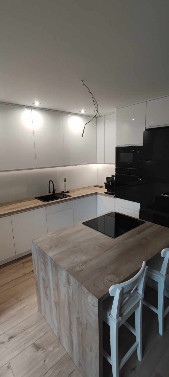 Nowoczesna kuchnia z białymi szafkami na wysoki połysk, drewnianym blatem i wyspą kuchenną z płytą indukcyjną; czarna armatura i sprzęt AGD, widoczne kable zwisające z sufitu.