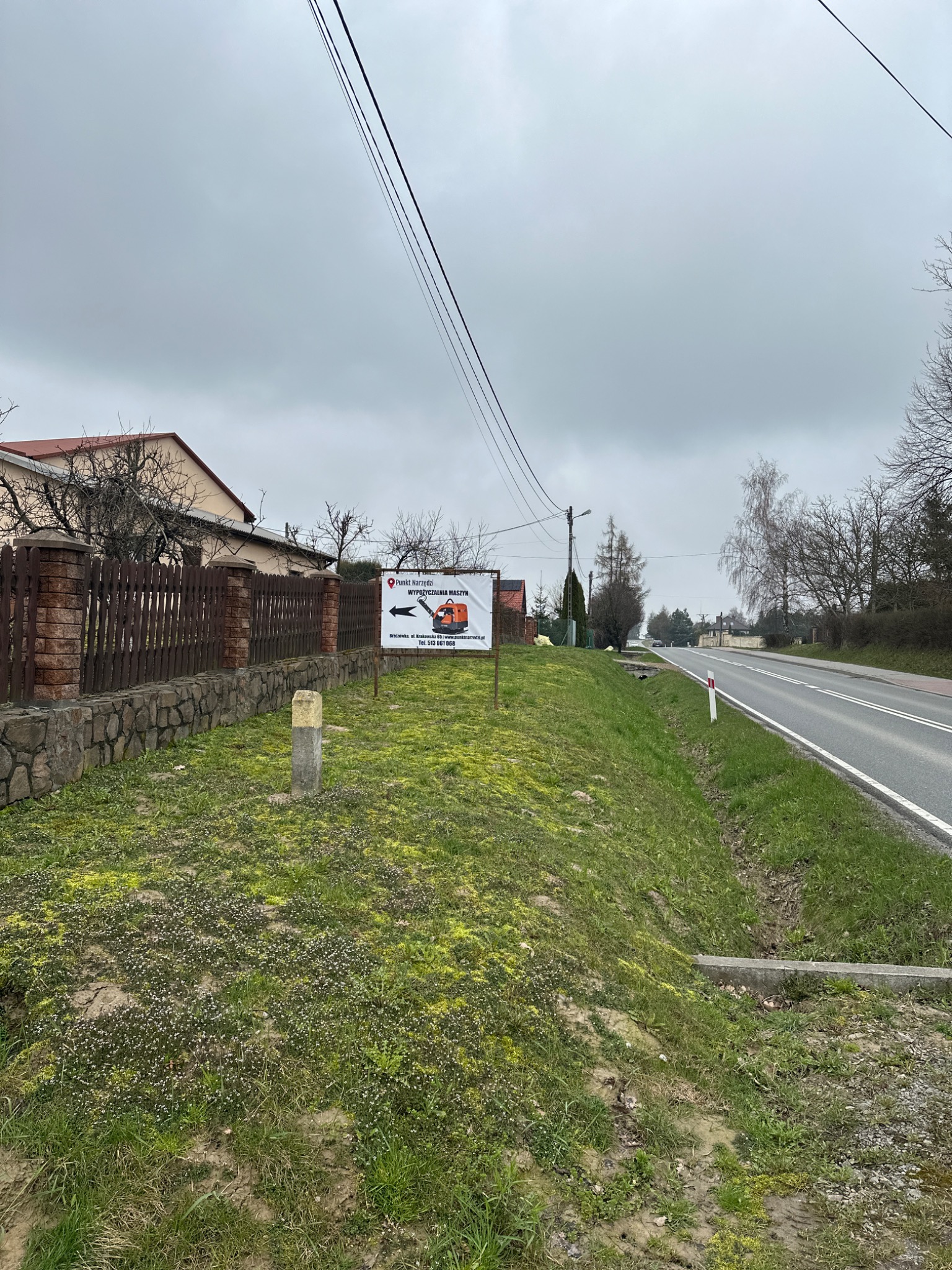 Tablica reklamowa z pomarańczową koparką i napisem 'Wypożyczalnia Maszyn' umieszczona przy drodze i trawniku, widok na budynek mieszkalny i słupy energetyczne.