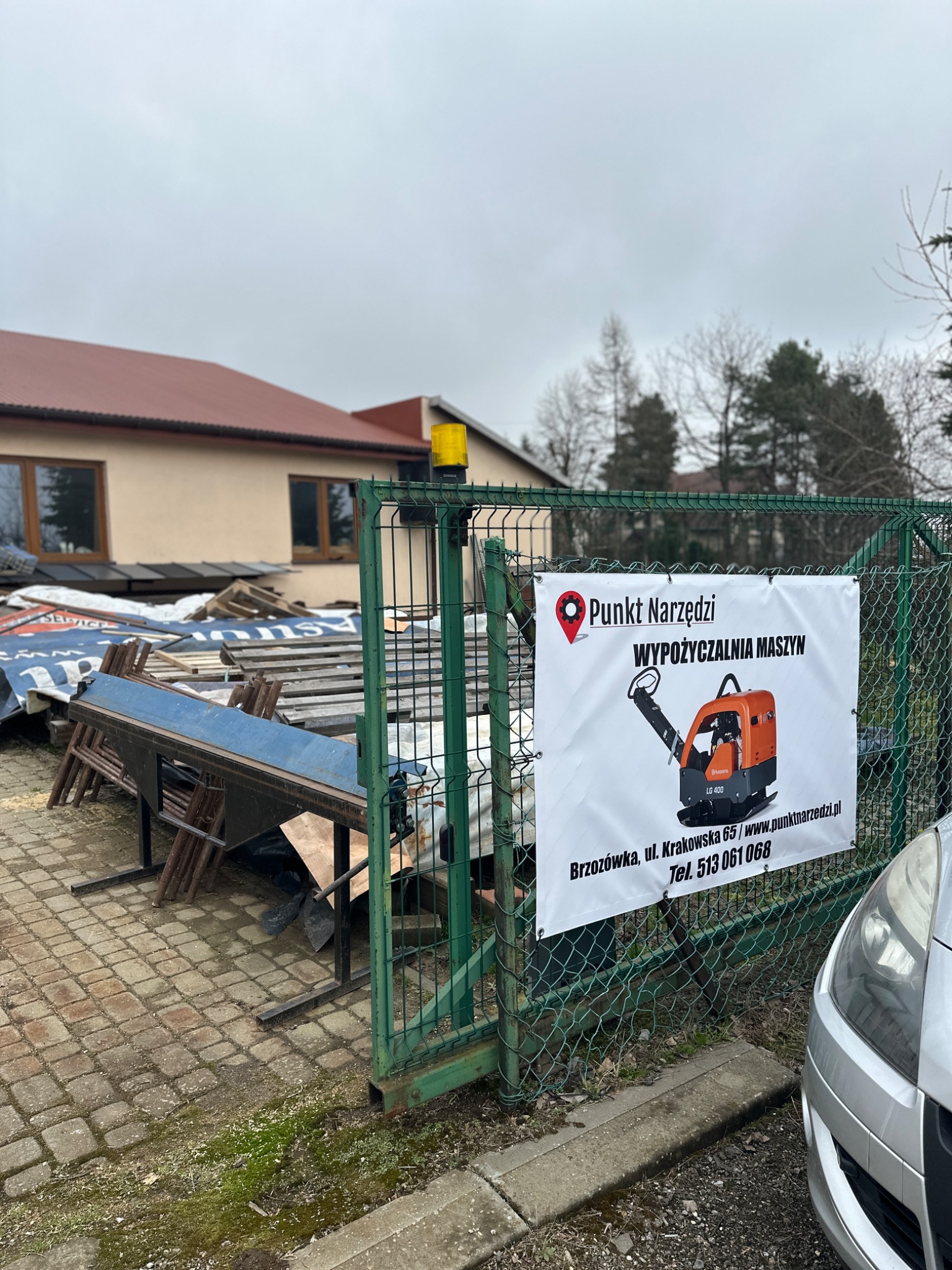 Baner reklamowy z ilustracją zagęszczarki gruntu, umieszczony na zielonym ogrodzeniu z siatki, w tle budynek i składowane materiały budowlane.