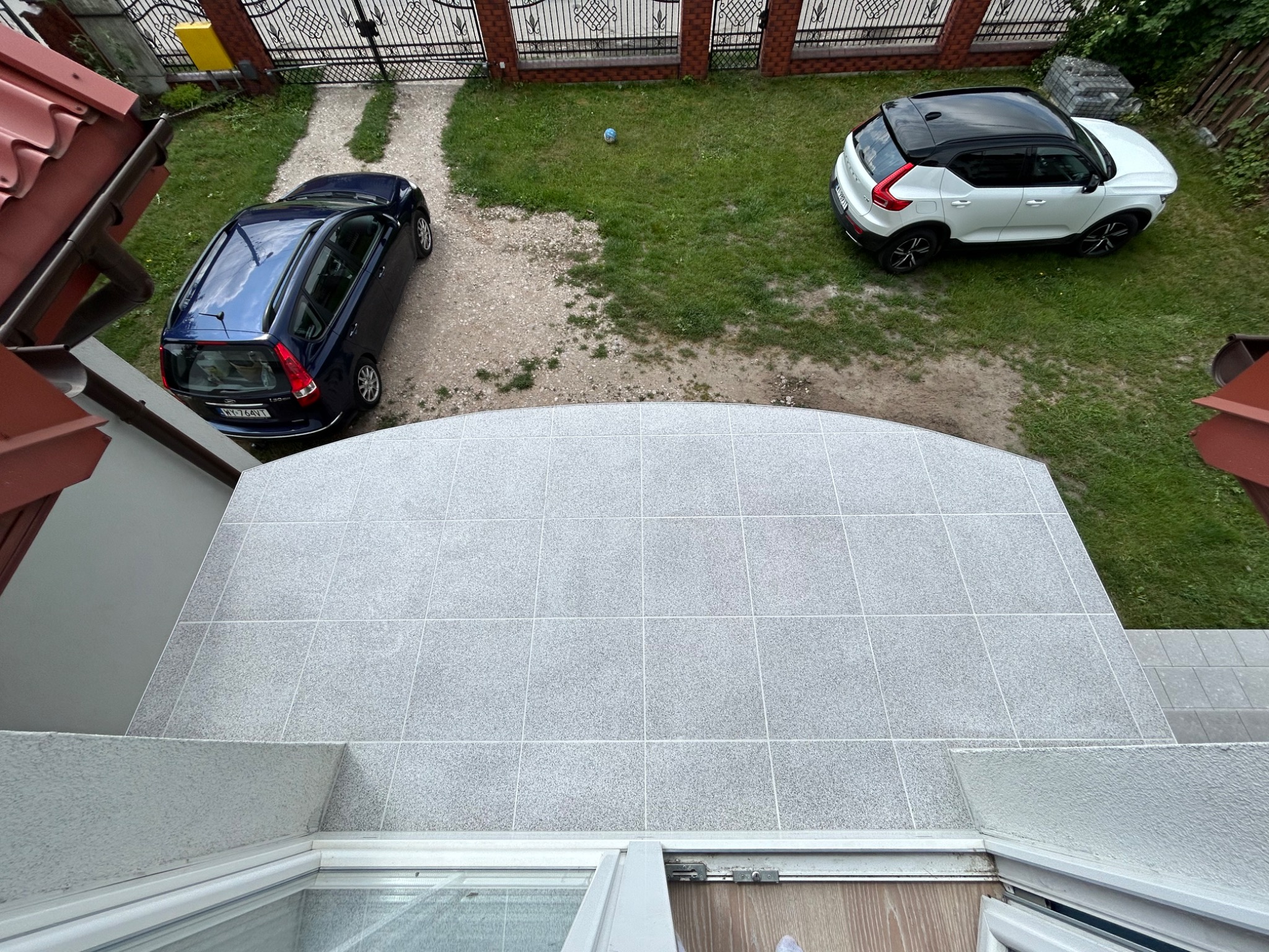 Zewnętrzny taras wyłożony szarymi płytkami, widok z góry, w tle trawnik, dwa samochody i ogrodzenie.