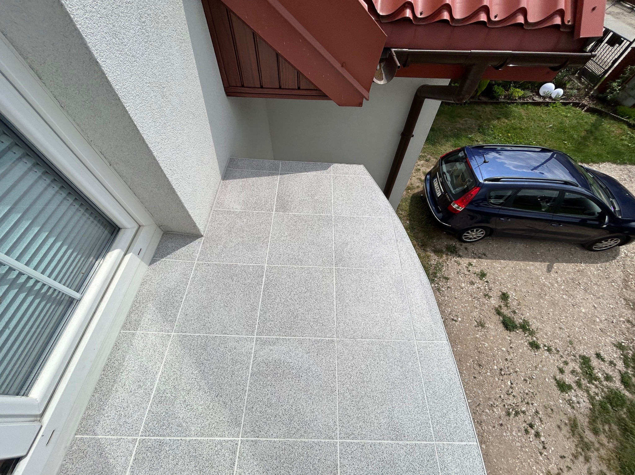 Wykończony balkon z szarymi płytkami, widok z góry, na zewnątrz zaparkowany granatowy samochód.