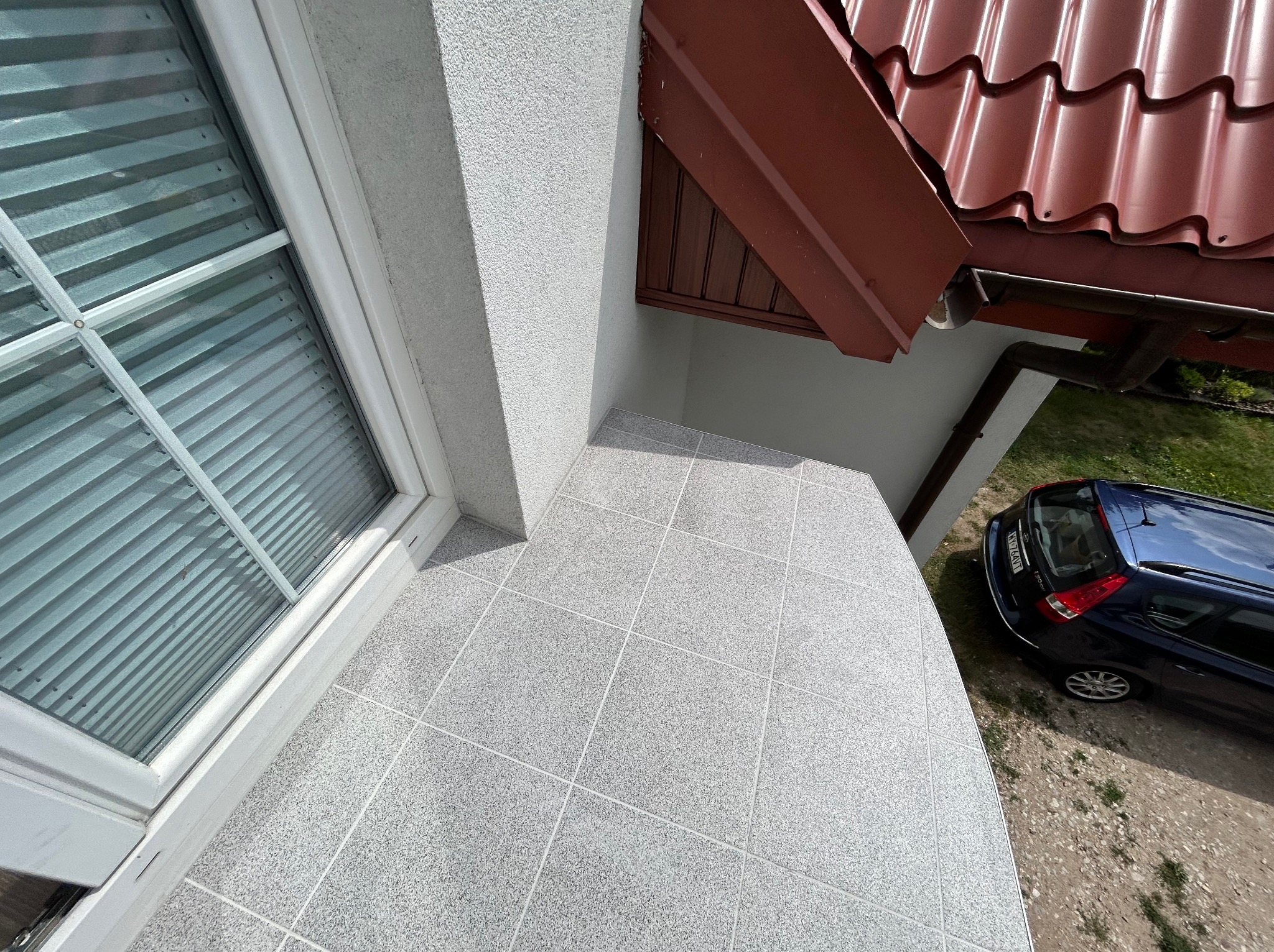 Wykończony balkon z szarą, nakrapianą płytką podłogową, widok z góry na fragment okna z żaluzjami, fragment dachu z brązową dachówką i zaparkowany samochód.