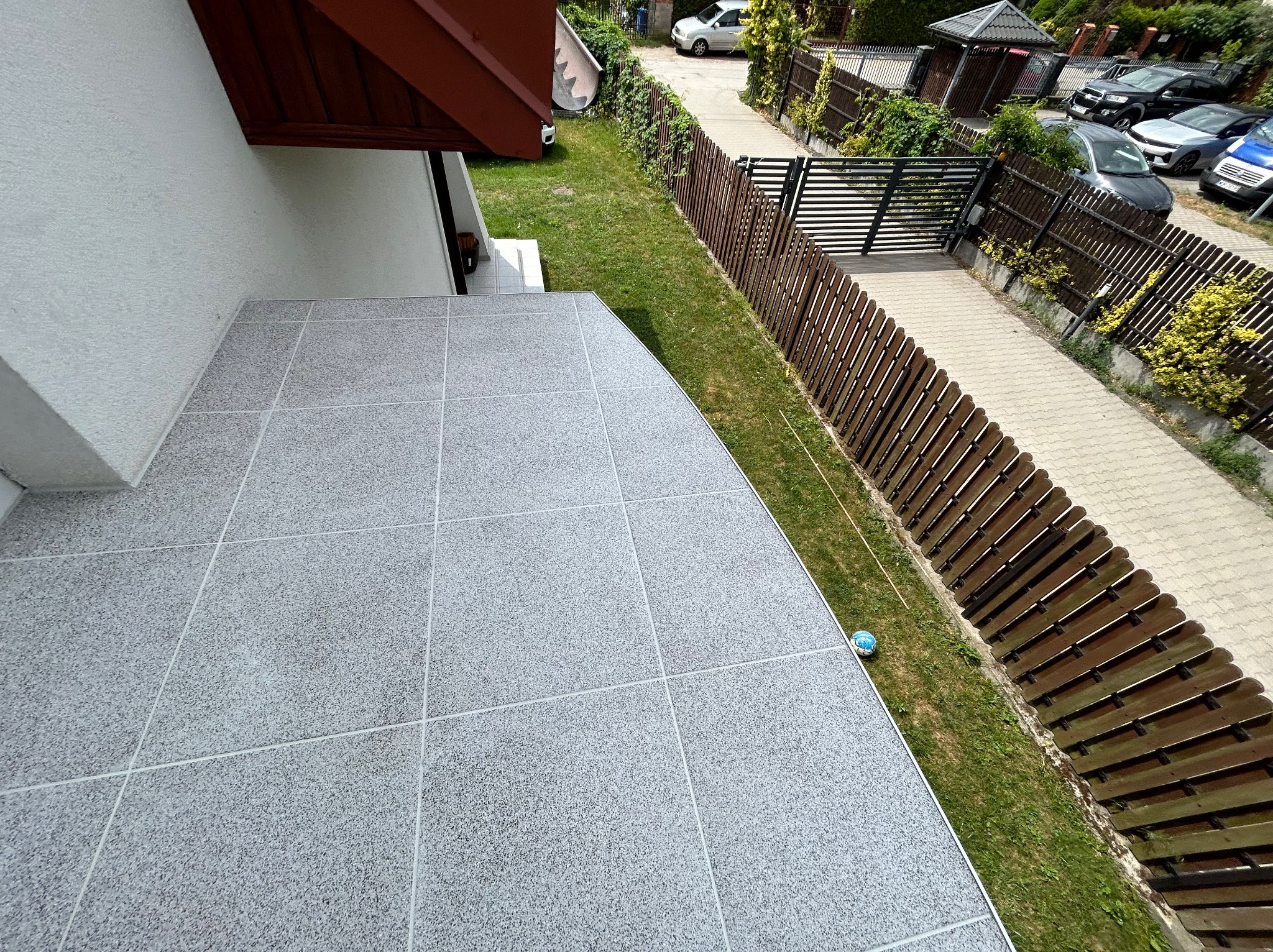 Wykończony balkon z szarymi płytkami o drobnej fakturze, widok z góry, z boku widoczny fragment trawnika i ogrodzenia, w rogu leży dziecięca piłka.
