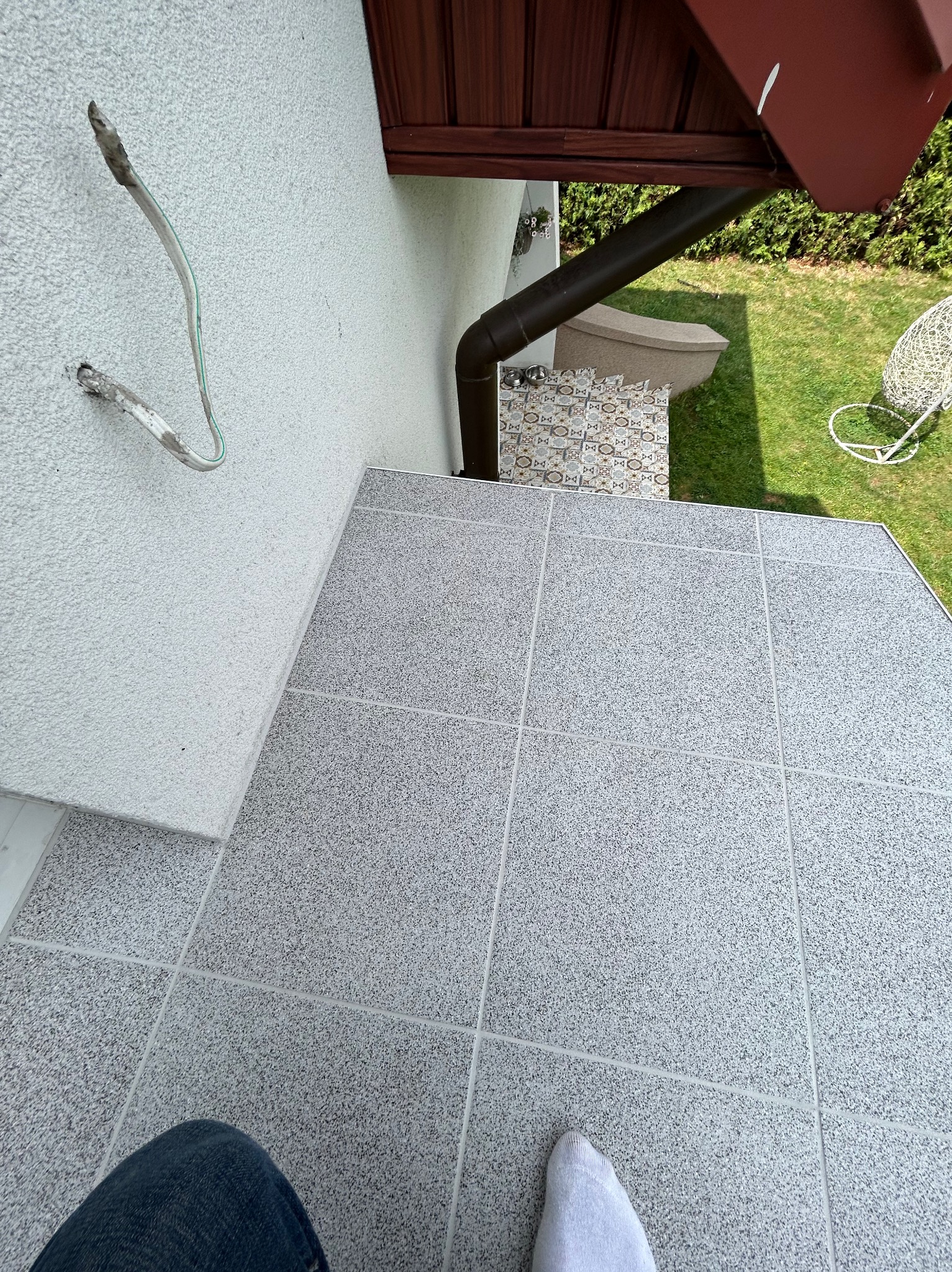 Szare płytki na zewnątrz budynku, widoczne wystające przewody elektryczne na ścianie, brązowa rynna i trawnik w tle.