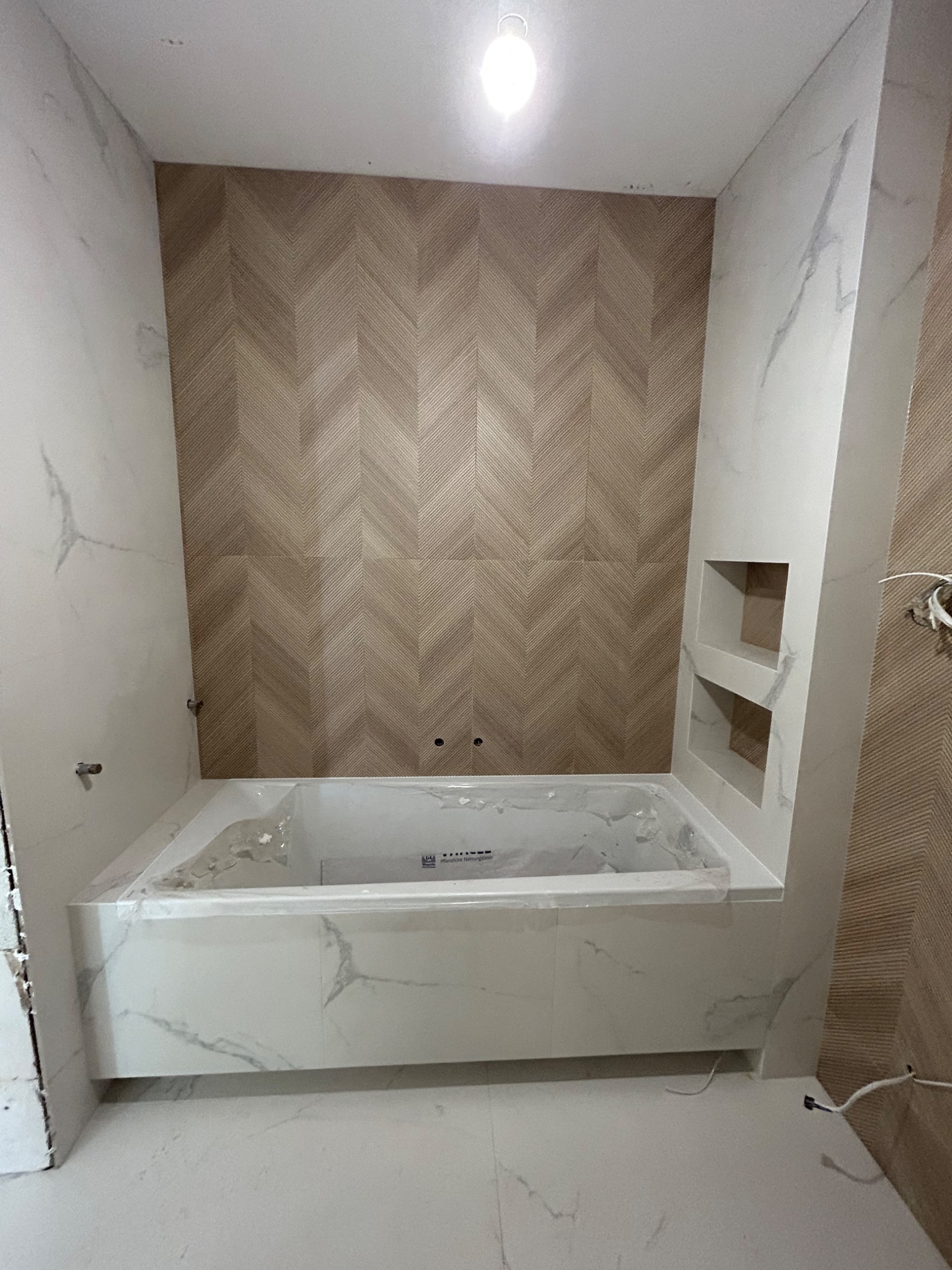 Wanna obudowana płytkami imitującymi marmur, z wnęką w ścianie również wykończoną płytkami; ściana wyłożona drewnopodobnymi płytkami w jodełkę, widoczne niedokończone prace instalacyjne.