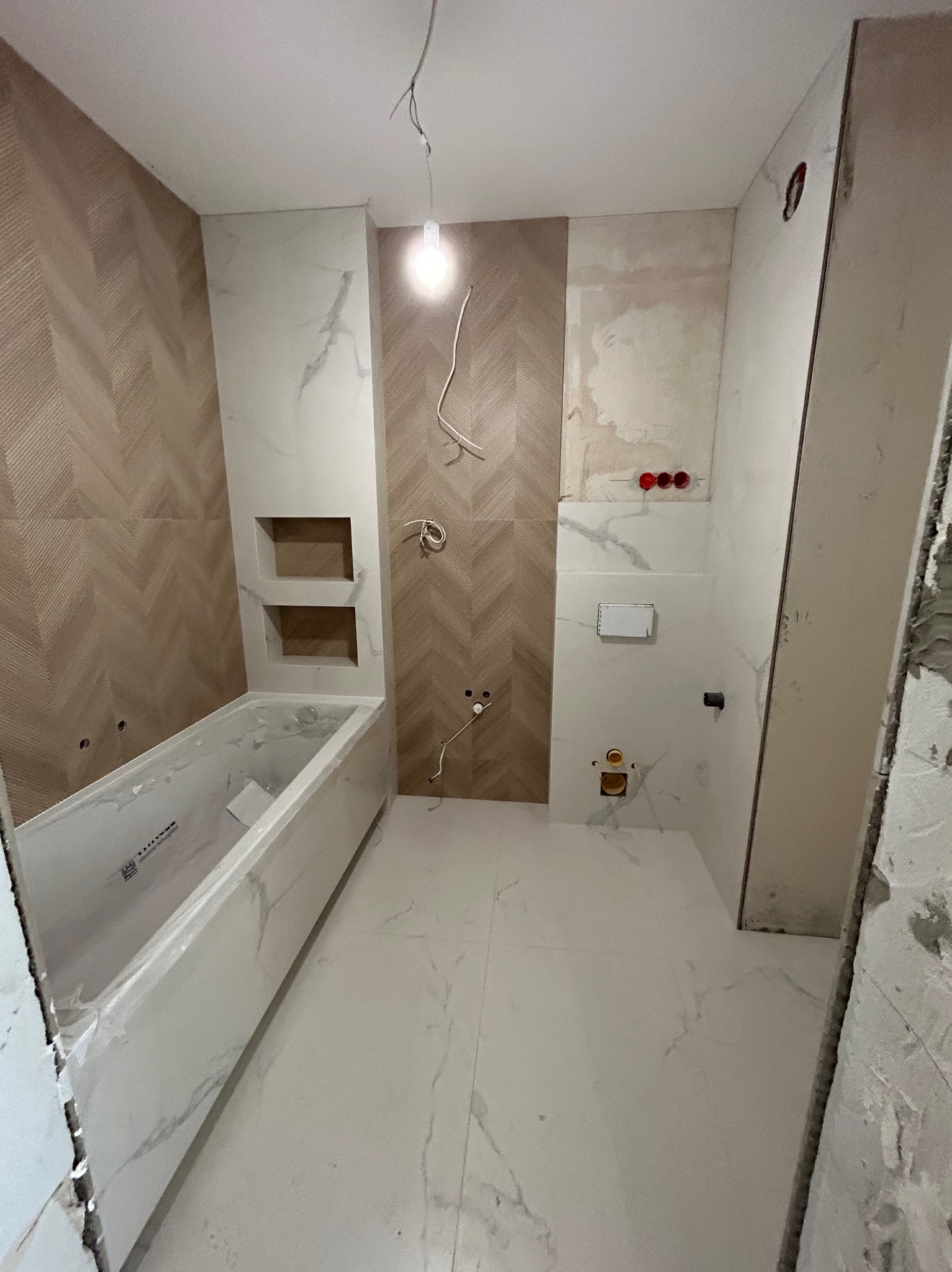 Wnętrze łazienki w trakcie prac wykończeniowych: położone płytki imitujące marmur na podłodze i częściowo na ścianach, z widoczną wanną obudowaną płytkami, instalacją elektryczną i hydrauliczną...