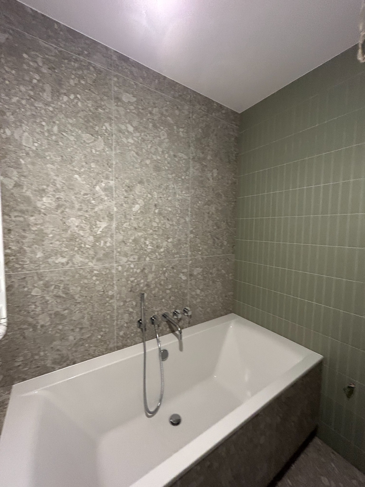 Nowoczesna łazienka z wanną obudowaną płytkami imitującymi beton, ściany wyłożone płytkami w dwóch odcieniach szarości i zieleni, bateria wannowa chromowana.