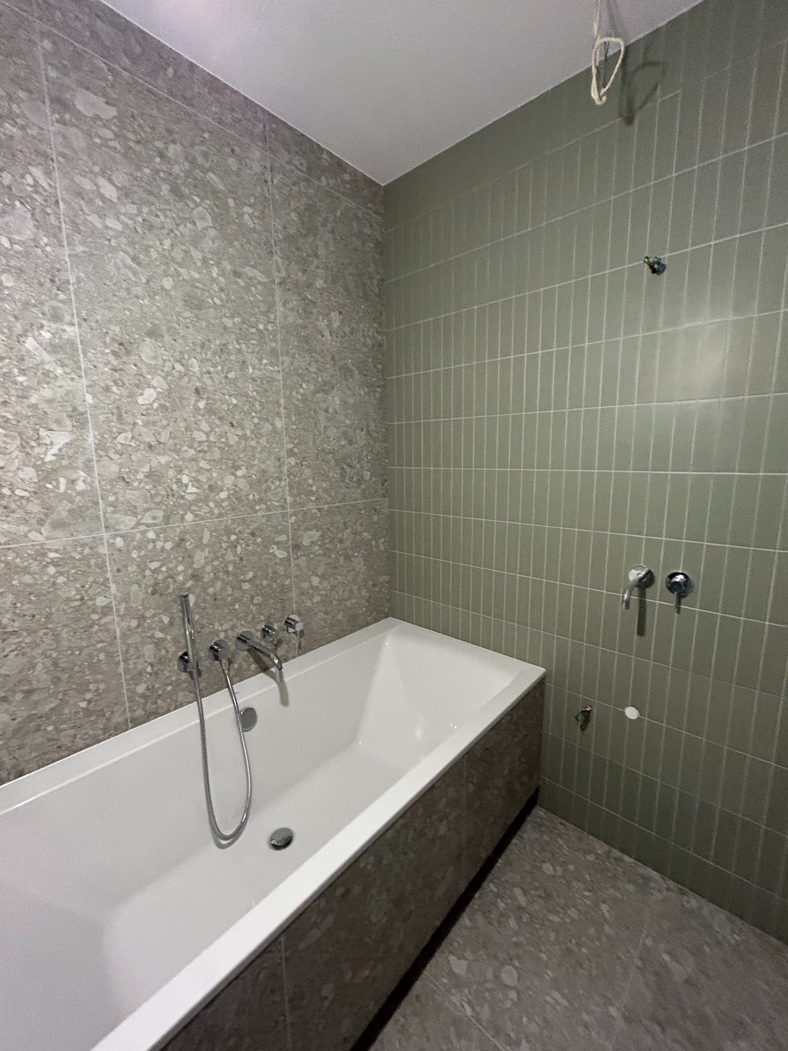 Wnętrze łazienki w trakcie prac wykończeniowych: biała wanna, ściana z szarych płytek imitujących kamień oraz ściana z pionowych, oliwkowych płytek, widoczne niedokończone instalacje wodne.