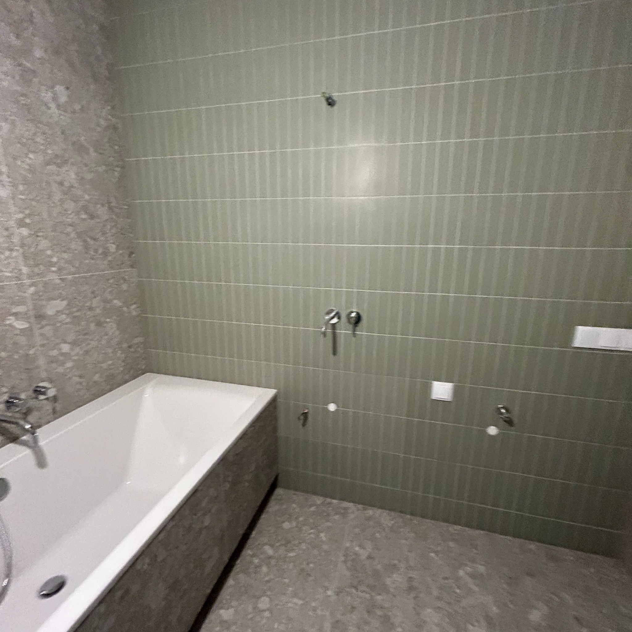 Wykończona łazienka z wanną, ściany wyłożone pionowymi, wąskimi, oliwkowymi płytkami oraz szarą, nakrapianą mozaiką. Widoczne baterie i przyłącza.
