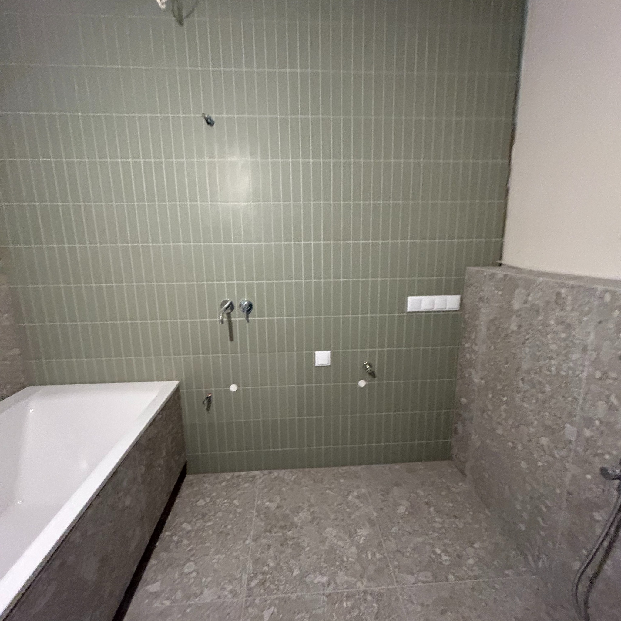 Wnętrze łazienki w trakcie wykańczania: zielone, wąskie płytki na ścianie w pionowym układzie, obudowana wanna z szarego lastryko, szara podłoga z lastryko, widoczne instalacje wodne, brak armatury.