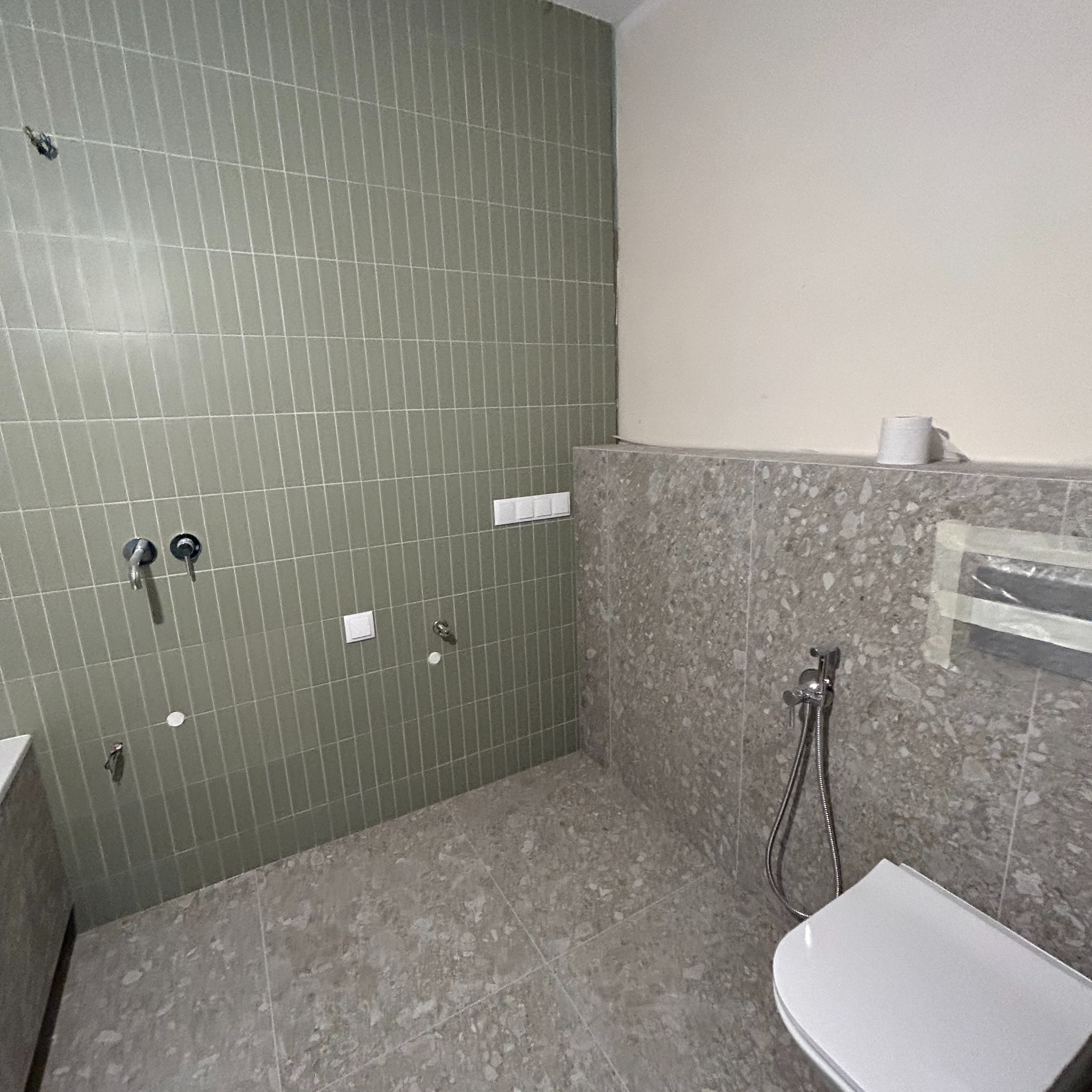 Łazienka w trakcie wykańczania: pionowo ułożone, oliwkowe płytki na ścianie, fragment ściany z szarym lastryko, zamontowane baterie, biała toaleta z bidetką, papier toaletowy na ścianie.