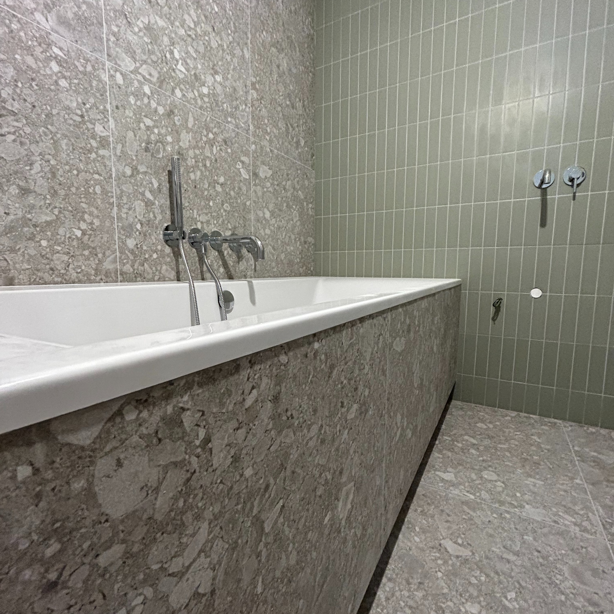 Nowoczesna łazienka z wanną obudowaną płytkami imitującymi lastryko, ściany wyłożone wąskimi, oliwkowymi płytkami.