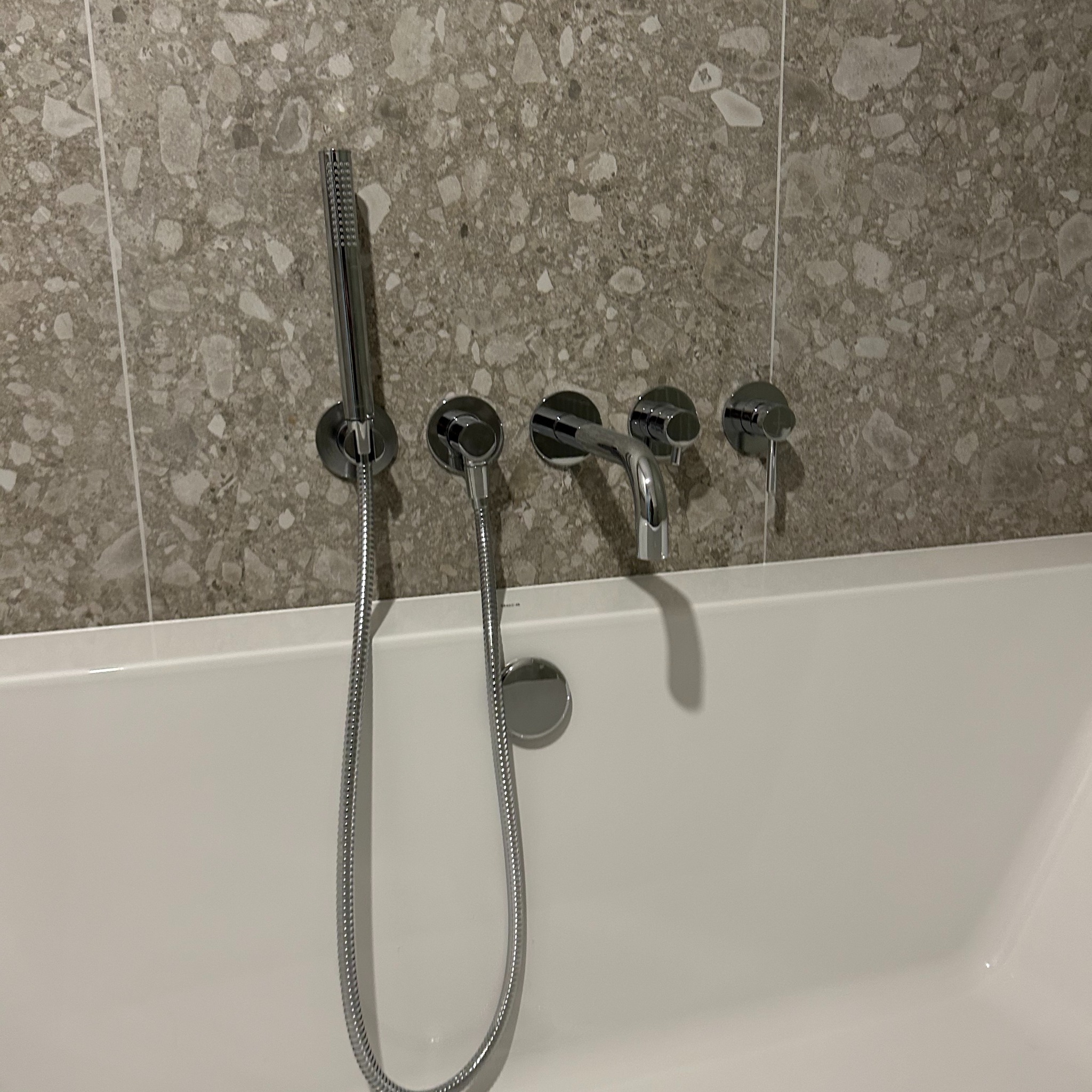 Bateria wannowa ścienna z chromowaną słuchawką prysznicową na tle ściany wykończonej płytkami imitującymi kamień.