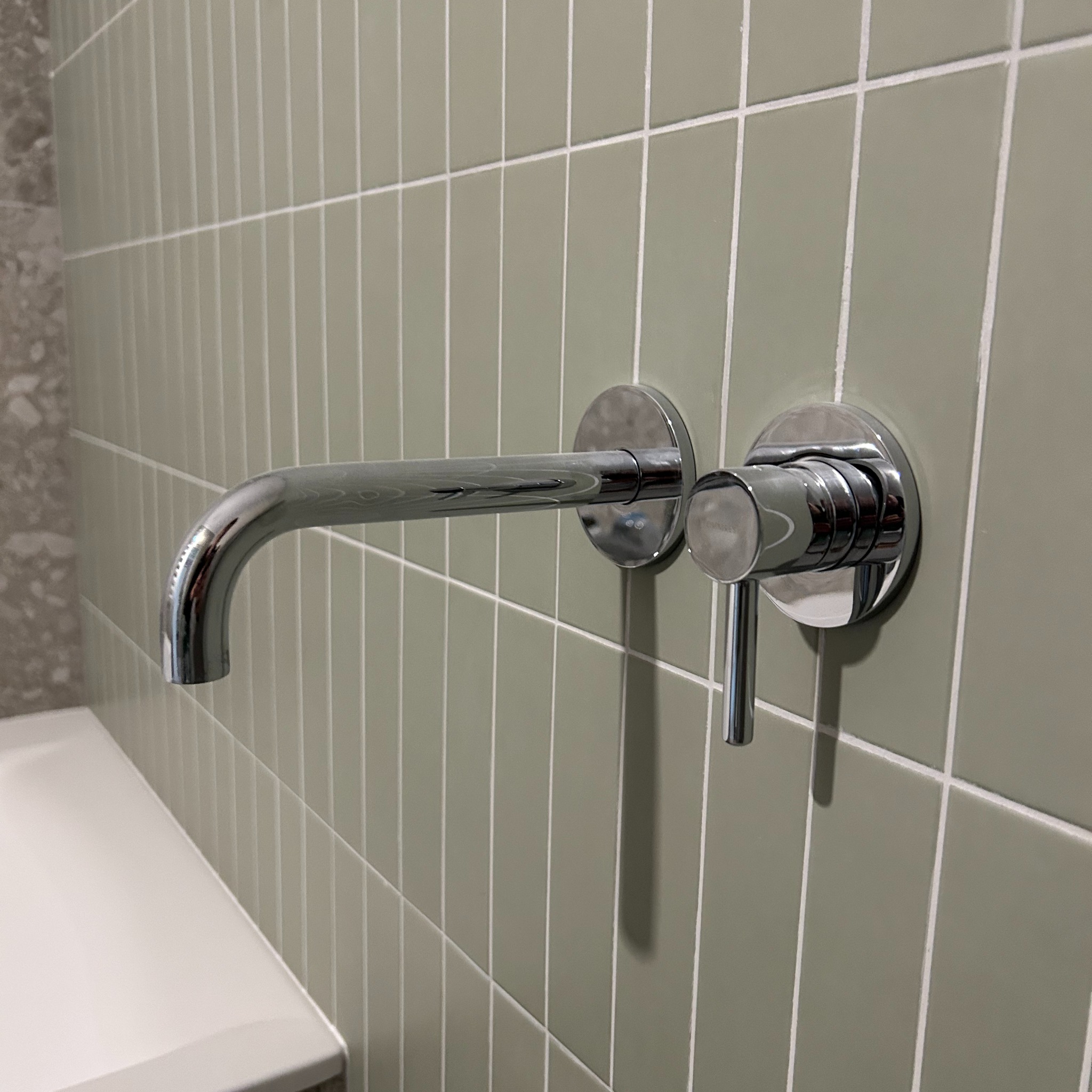 Bateria umywalkowa chromowana, zamontowana na ścianie z podłużnych, oliwkowych płytek ceramicznych w łazience, widok z boku.