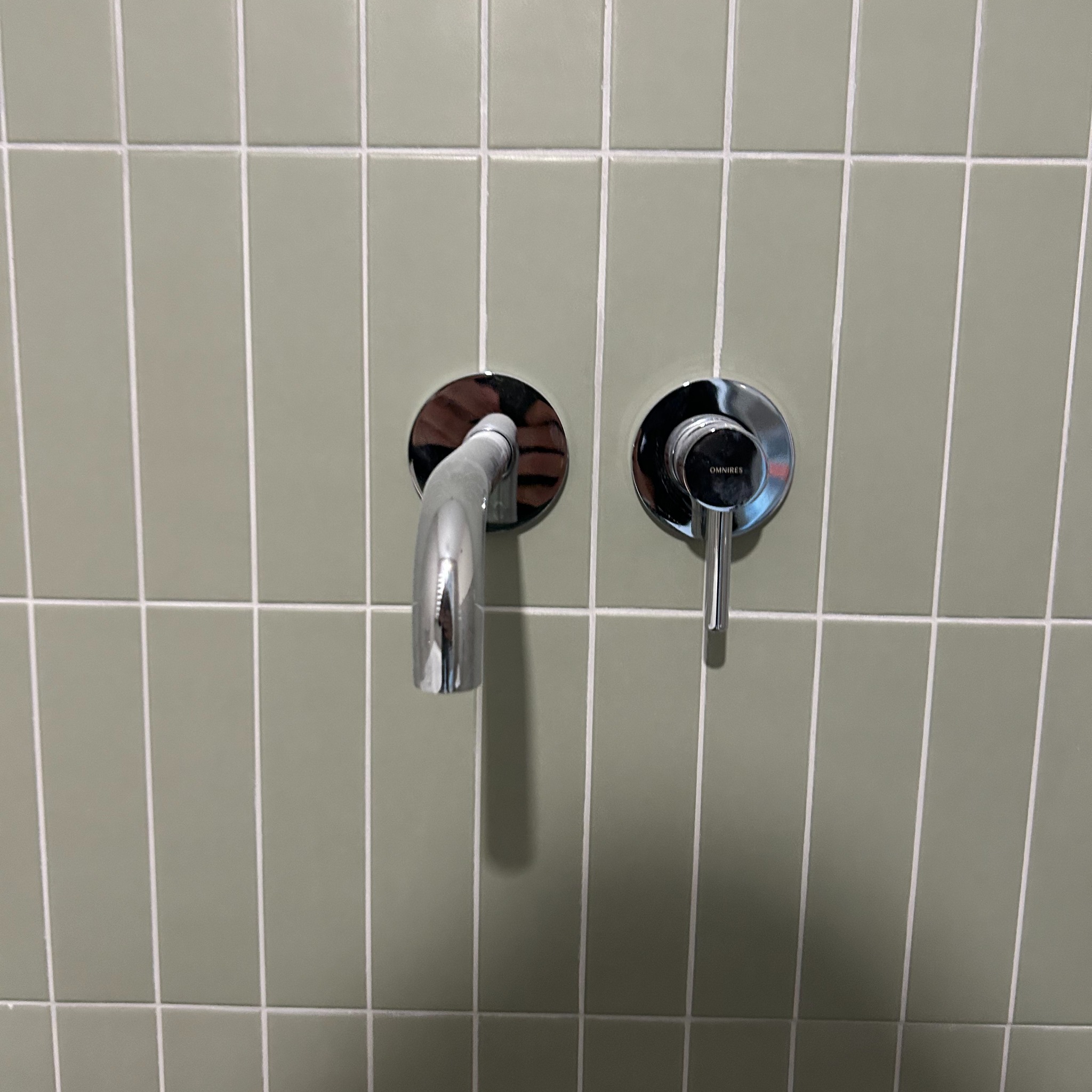 Bateria umywalkowa chromowana, zamontowana na ścianie z pionowych, oliwkowozielonych płytek z jasną fugą.