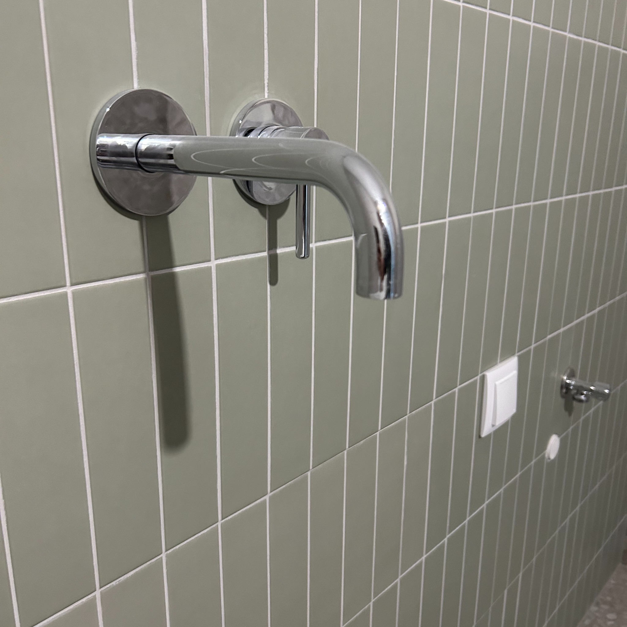 Bateria umywalkowa chromowana, zamontowana na ścianie wyłożonej pionowymi, oliwkowymi płytkami z białymi fugami, obok włącznik światła i uchwyt na papier toaletowy.