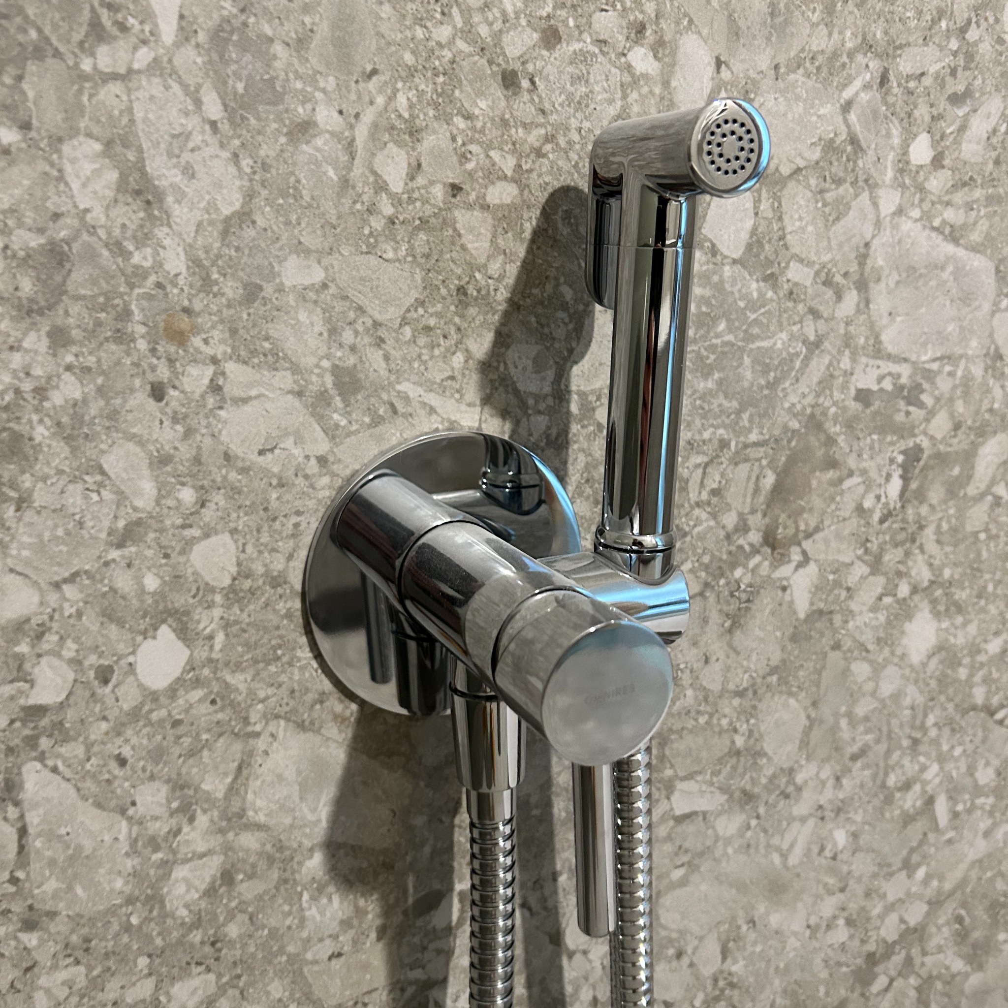 Bidetta chromowana, zamontowana na ścianie z szarego lastryko, widoczne wężyki i dysza.