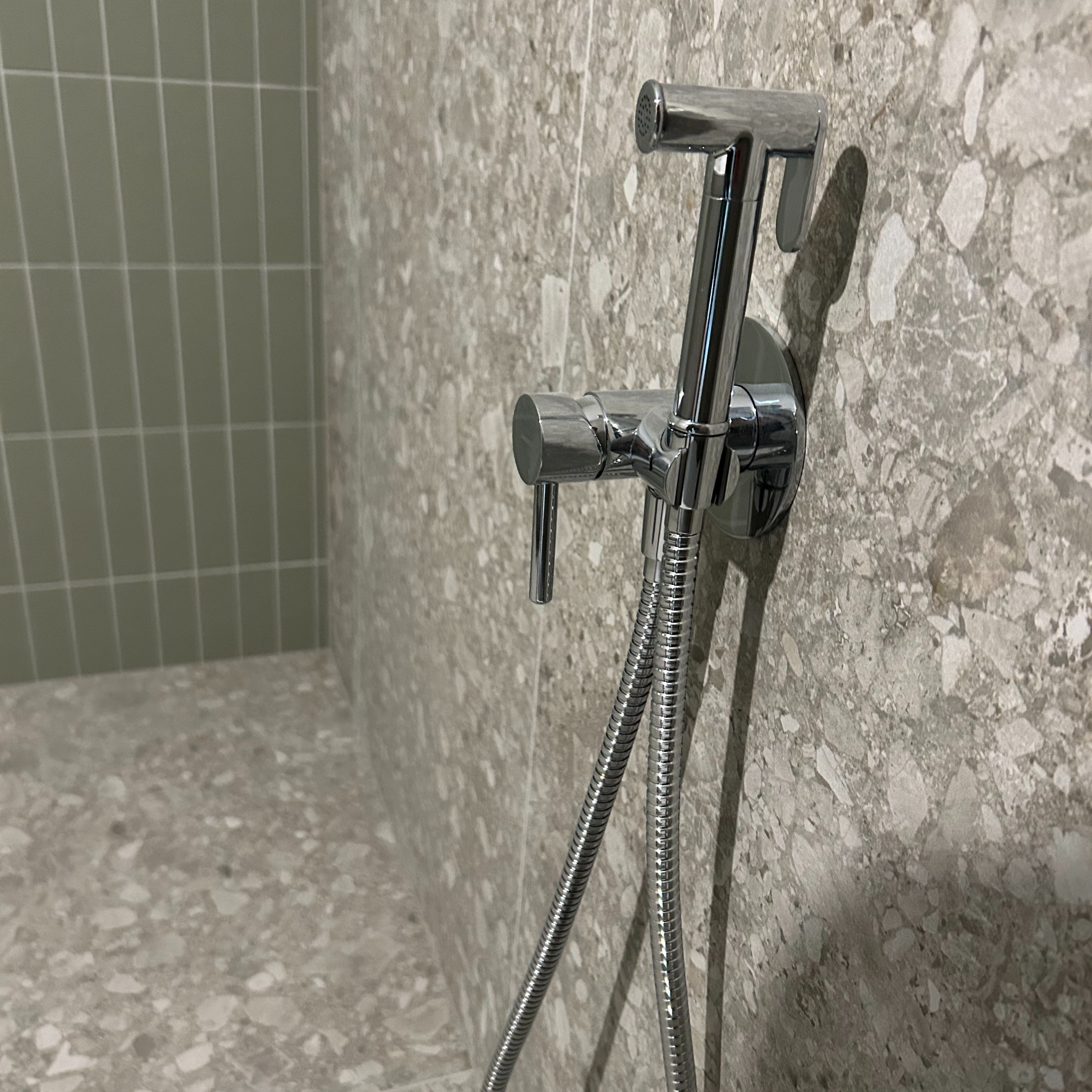 Bidetta chromowana z wężem na tle ściany wyłożonej płytkami imitującymi lastryko i fragmentem ściany z zielonymi, podłużnymi płytkami.