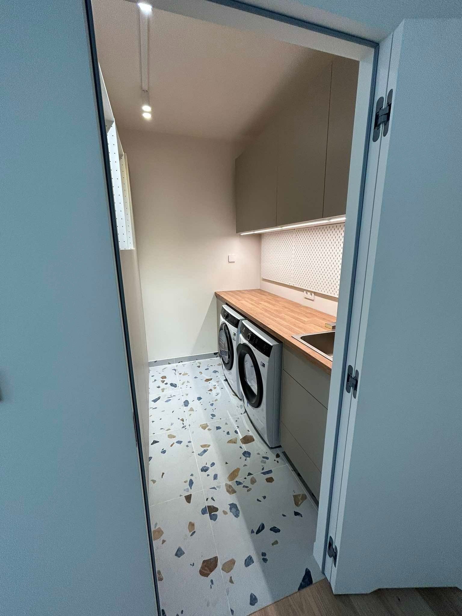 Nowoczesna pralnia z dwoma pralkami, drewnianym blatem, szafkami w kolorze beżowym, podświetleniem LED podszafkowym i podłogą w stylu lastryko, widziana zza uchylonych drzwi.