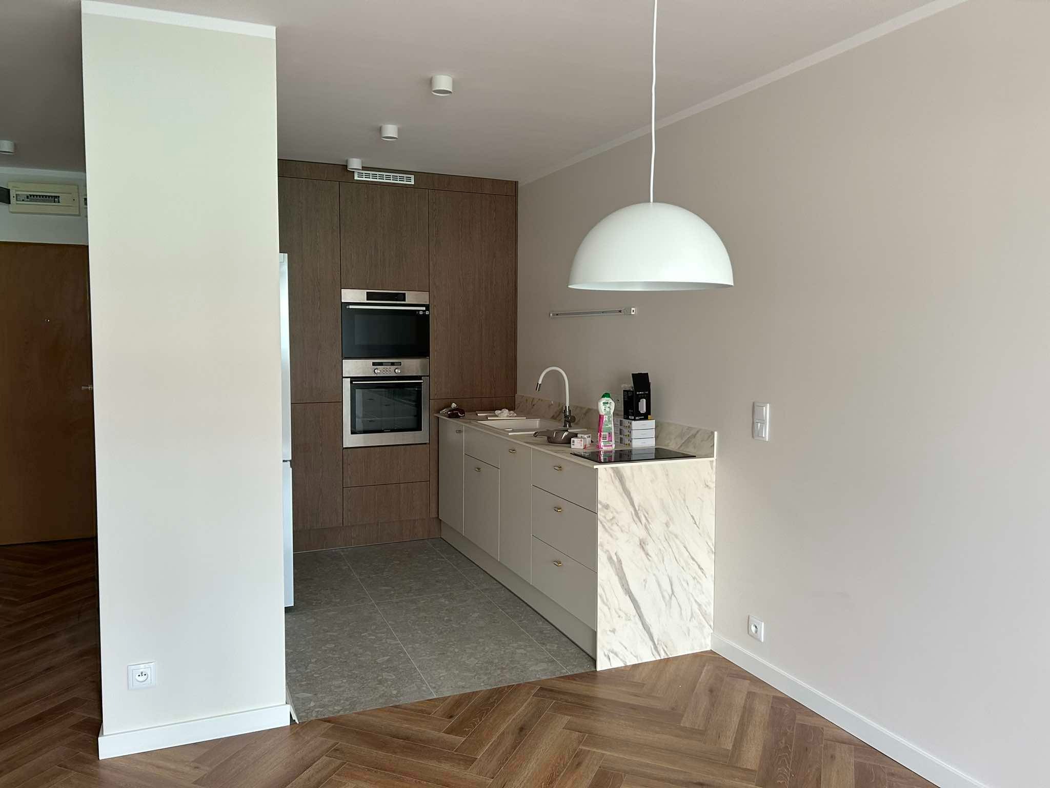 Jasna kuchnia z drewnianą zabudową, marmurowym blatem i parkietem ułożonym w jodełkę. Widoczne sprzęty AGD oraz oświetlenie sufitowe.