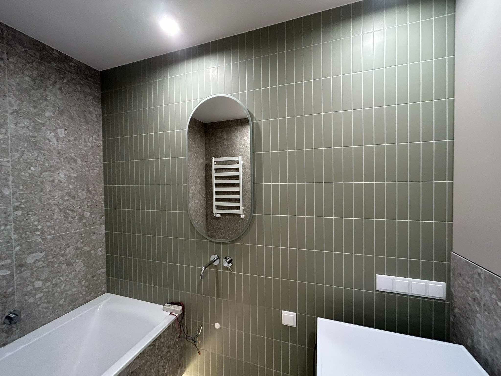 Nowoczesna łazienka z pionowo ułożonymi, oliwkowymi płytkami, owalnym lustrem odbijającym grzejnik drabinkowy i fragmentem ściany wykończonej płytami imitującymi szary kamień.