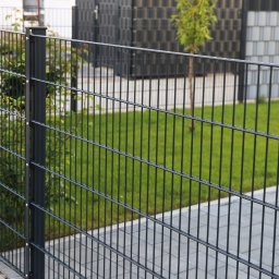 Płot-Bruk Tomasz Piskorski - Nowoczesne, metalowe ogrodzenie panelowe w kolorze grafitowym, widoczne z bliska, oddzielające posesję z zielonym trawnikiem i młodym drzewkiem od chodnika wyłożonego szarą kostką brukową.