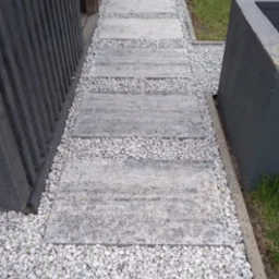 Ścieżka ogrodowa z szarych płyt betonowych, wypełniona białym grysem, prowadząca wzdłuż ciemnej, drewnianej konstrukcji.