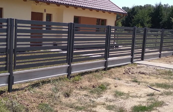 Nowoczesne, poziome przęsła w grafitowym odcieniu, tworzące ogrodzenie na betonowej podmurówce, z widocznym domem jednorodzinnym w tle.
