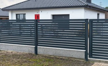Nowoczesne ogrodzenie panelowe w kolorze antracytowym z poziomymi listwami, zamontowane na niskiej, betonowej podmurówce. W tle widoczny biały dom z czerwonymi drzwiami wejściowymi i szarą dachówką.