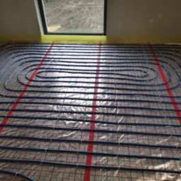 Niebieskie rury układane spiralnie na folii z czerwoną kratką, przygotowanie do wylewki ogrzewania podłogowego w jasnym pomieszczeniu z oknem dachowym.