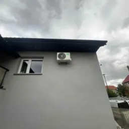 Biała jednostka zewnętrzna klimatyzacji zamontowana na szarej ścianie budynku, widoczna z dołu pod pochmurnym niebem. Obok okno z białymi ramami.