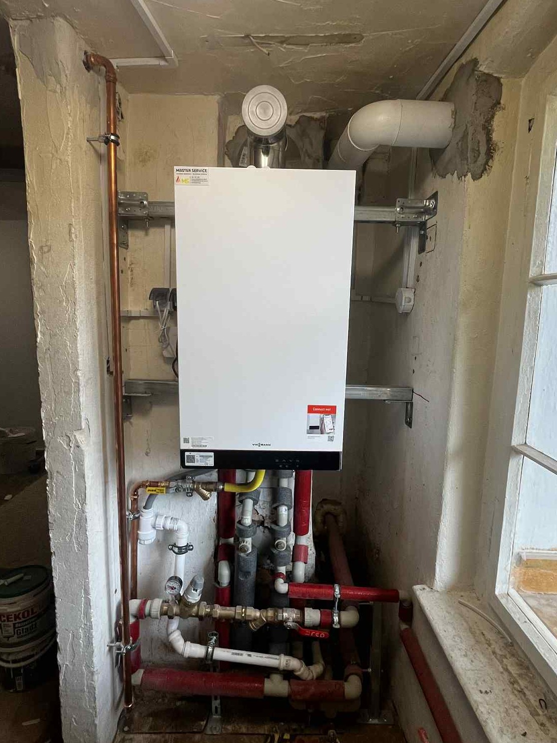 Biały piec gazowy Viessmann zamontowany na ścianie z instalacją hydrauliczną i rurami. Widoczne elementy montażowe i fragment okna. Ściany w surowym stanie.