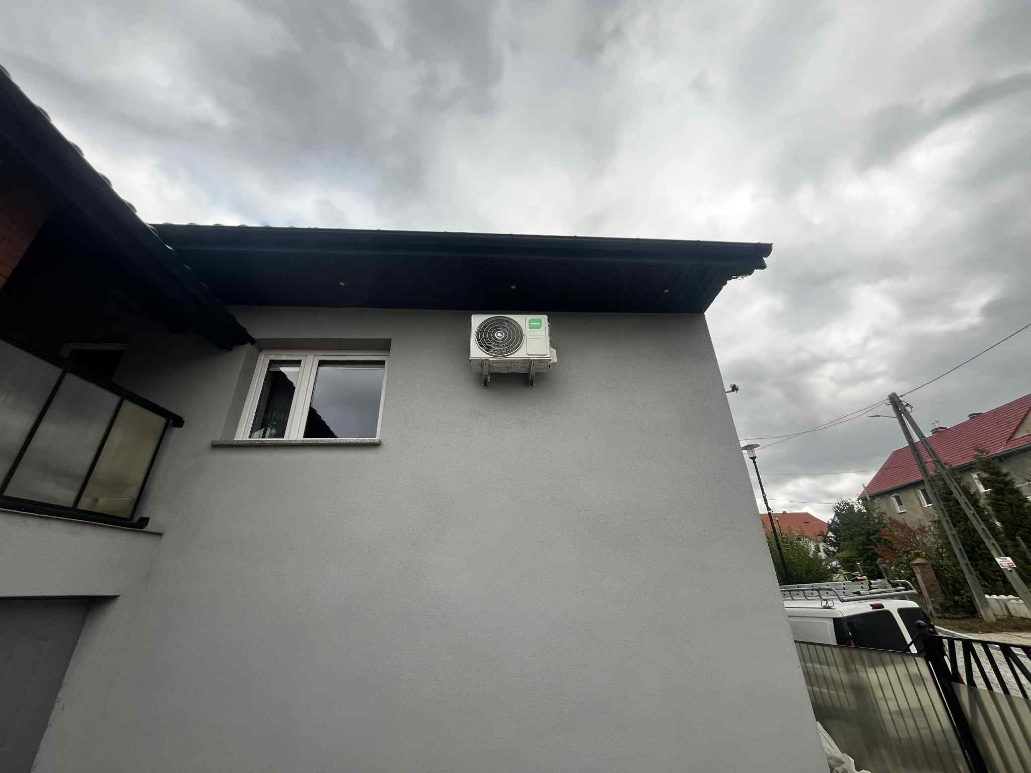 Biała jednostka zewnętrzna klimatyzacji zamontowana na szarej ścianie budynku, widoczna z dołu pod pochmurnym niebem. Obok okno z białymi ramami.